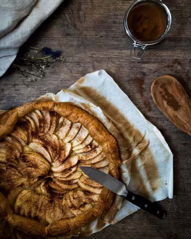 A cinnamon coated apple pie sliced on parchment paper with a knife on a wooden table