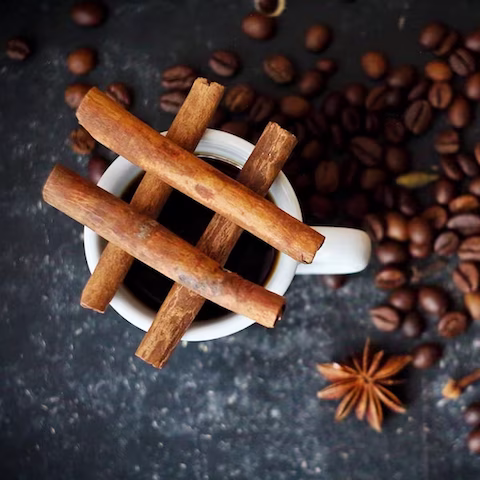 Four cinnamon sticks placed on the rim of a cup, surrounded by coffee beans on a dark surface