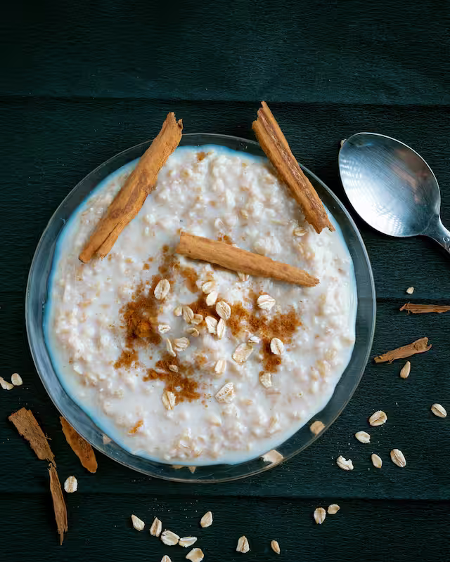 A bowl of creamy oatmeal topped with cinnamon sticks and sprinkled oats on a dark surface