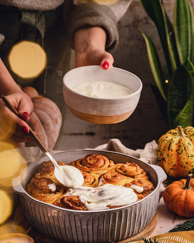 Freshly baked cinnamon rolls in a dish being drizzled with icing beside a bowl